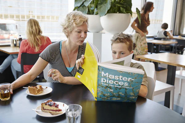 Een moeder en haar zoontje genieten in het museumcafé van frisdrank en taart. Het zoontje bladert door het boek Meesterwerken 